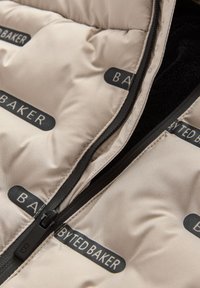 Beige vuorillinen takki mustalla vetoketjulla, jossa on toistuvasti "BAKER"-logoja. Sisävuori on teksturoitu ja tumma, mikä luo kontrastin ulkopinnan kanssa.