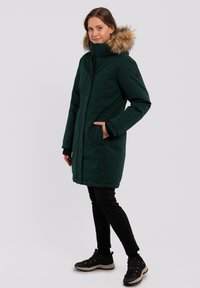 Parka verde con cappuccio rifinito in pelliccia, zip anteriore, grandi tasche e lunghezza fino a metà coscia, abbinata a pantaloni neri e calzature.