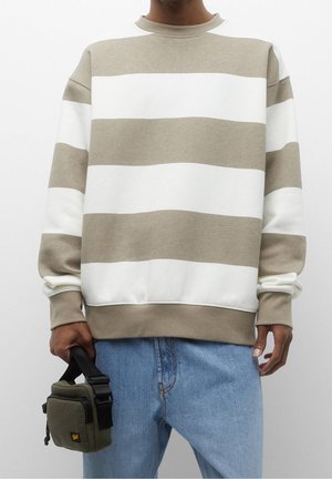 Torso de persona con sudadera de rayas anchas beige y blanca y vaqueros azules claros, sosteniendo un bolso pequeño verde oliva con correa negra.