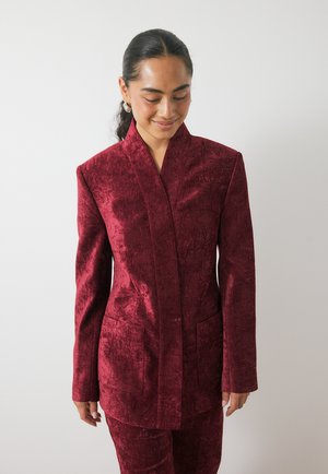 Blazer ajustado burdeos con un acabado de terciopelo texturizado, que presenta un cuello alto y dos bolsillos frontales. Silueta elegante y moderna.