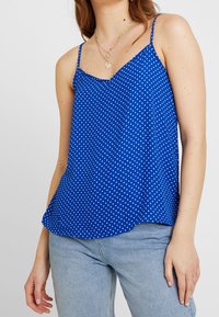 Žena v bezrámovém modrém topu s puntíky a světle modrých džínách, doplněná o vrstvené zlaté náhrdelníky na jednobarevném pozadí.