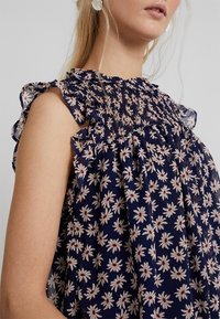 Top floreale navy con motivi di margherite bianche e rosse; corpetto arricciato; dettagli arricciati sulle spalle; tessuto leggero; vestibilità ampia.