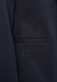 Ύφασμα σε ναυτικό μπλε με διακριτικό σχέδιο ψαροκόκκαλο και οριζόντια τσέπη με επιστόμιο σε ένα ραμμένο ένδυμα.