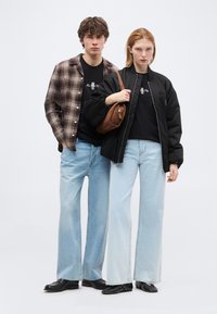 Νεαρός άνδρας και γυναίκα όρθιοι, και οι δύο φορούν ανοιχτό μπλε τζιν με φαρδιά πόδια και μαύρα τοπ, με τη γυναίκα να κρατά καφέ τσάντα ώμου.