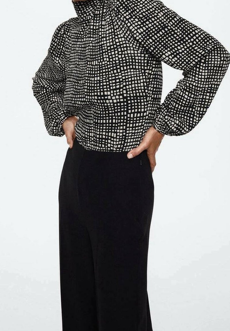 Personne portant un chemisier à manches longues à carreaux noir et blanc, rentré dans un pantalon noir taille haute, les mains sur les hanches.