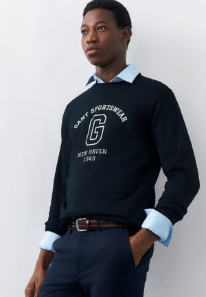 Mladý muž stojí, oblečený v čiernej mikine s nápisom „GANT SPORTSWEAR NEW HAVEN 1949“ cez svetlomodrú košeľu s golierom, ruky v džepoch.