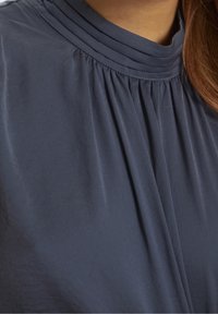 Námořnická modrá blůza s plisovaným designem, vysokým límcem a měkkou, lehkou tkaninou pro hladkou texturu a splývavost.