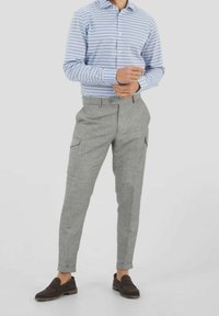 Man in een lichtblauw en wit gestreept overhemd met lange mouwen, grijze broek met opgerolde pijpen en cargozakken, en donkerbruine instappers staat de manchetten aan te passen.