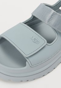 Lyseblå sandal med en glatt gummioverdel, justerbar stropp og en teksturert fotseng med en tykk såle. UGG-logo preget på siden.