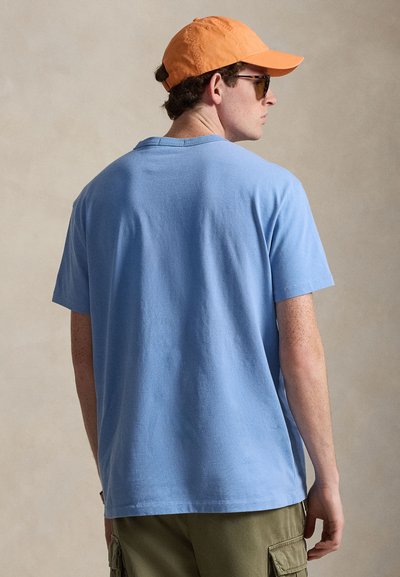 Άνδρας με πορτοκαλί καπέλο, γυαλιά ηλίου, ανοιχτό μπλε μπλουζάκι και ελαιδί παντελόνι cargo, στέκεται γυρισμένος με την πλάτη προς ένα απλό φόντο.