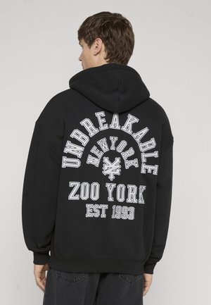 Ung mand set bagfra, iført en sort hættetrøje med hvid tekst på ryggen, der lyder "UNBREAKABLE NEW YORK ZOO YORK EST 1993".