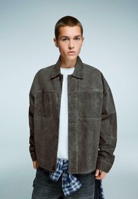 Siva overshirt jakna s ovratnikom, sprednjimi žepi in sproščenim krojem, oblečena preko bele srajce, z vezanim flanelom okoli pasu.