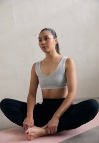 Gray cropped tank top, black loose pants, sitting cross-legged on a pink yoga mat, with a plain light backdrop and a relaxed posture.