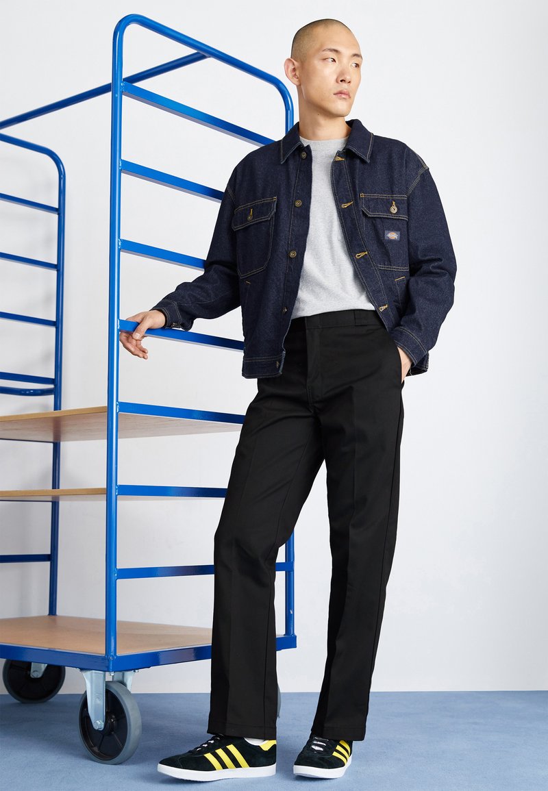 Denim jacket with a relaxed fit, gray t-shirt, black trousers, and black suede sneakers with yellow stripes. Blue clothing rack in background.