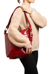 Borsa a spalla in pelle rossa con finitura liscia, caratterizzata da una tracolla larga e un dettaglio in oro. Indossata di lato contro un maglione beige.