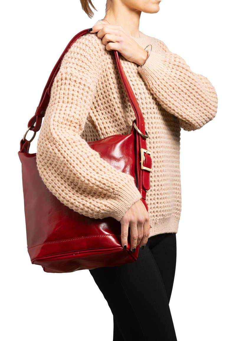 Borsa a spalla in pelle rossa con finitura liscia, caratterizzata da una tracolla larga e un dettaglio in oro. Indossata di lato contro un maglione beige.