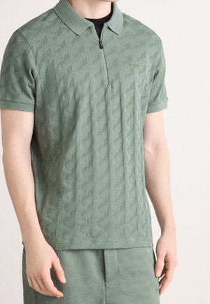 Mężczyzna ubrany w jasnzie zieloną teksturowaną koszulkę polo z krótkim rękawem i suwakiem oraz pasujące szorty, stojący na tle jednolitego jasnego tła.