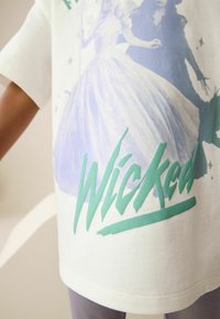 Biała koszulka z krótkim rękawem z nadrukowanym wzorem na jasnoniebieskim tle, przedstawiająca postacie i słowo "Wicked" w zielonym kolorze.