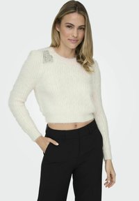White cropped sweater with fuzzy texture, ribbed detailing, and embellished shoulder accents, paired with black tailored pants.