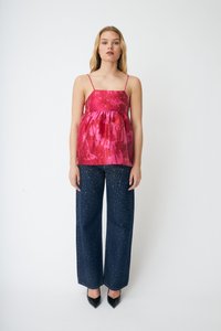 Ung kvinne iført ermeløs rosa mønstret topp, vide mørke jeans med små hvite prikker, og svarte spisse hæler, stående mot hvit bakgrunn.