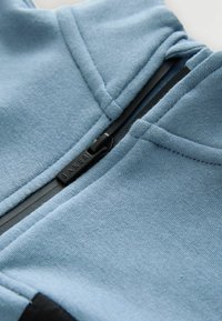 Jasnoniebieska bluza z kapturem na zamek błyskawiczny, wykonana z fakturowanej tkaniny, z czarnym zamkiem błyskawicznym i etykietą "BAKER", oraz kontrastującymi czarnymi detalami na dole.