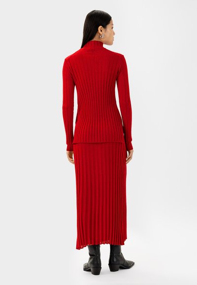 Rotes geripptes Strickkleid mit langen Ärmeln und hohem Kragen, das eine körperbetonte Silhouette und einen ausgestellten Saum bietet. Kombiniert mit schwarzen Stiefeletten.