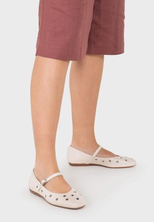 Nogi w mauve krótkich spodenkach do kolan i białych balerinach Mary Jane z srebrnymi ćwiekami na jednolitym tle.