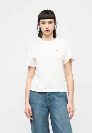 Nuori nainen, jolla on lyhyet tummat hiukset, pukeutuneena yksinkertaiseen valkoiseen t-paitaan ja väljään sinisiin farkkuihin, seisoo vaaleanharmaan taustan edessä.