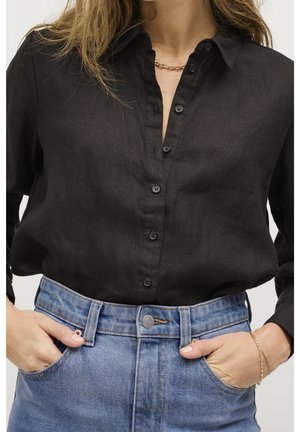 Femme portant une chemise noire boutonnée rentrée dans un jean bleu taille haute, les mains dans les poches avant, avec un collier et un bracelet en chaîne dorée.