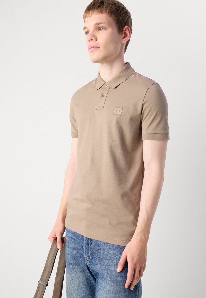 Polo shirt beige en coton, avec un col classique, une patte de boutonnage à deux boutons et un petit logo. Porté avec un jean en denim bleu.