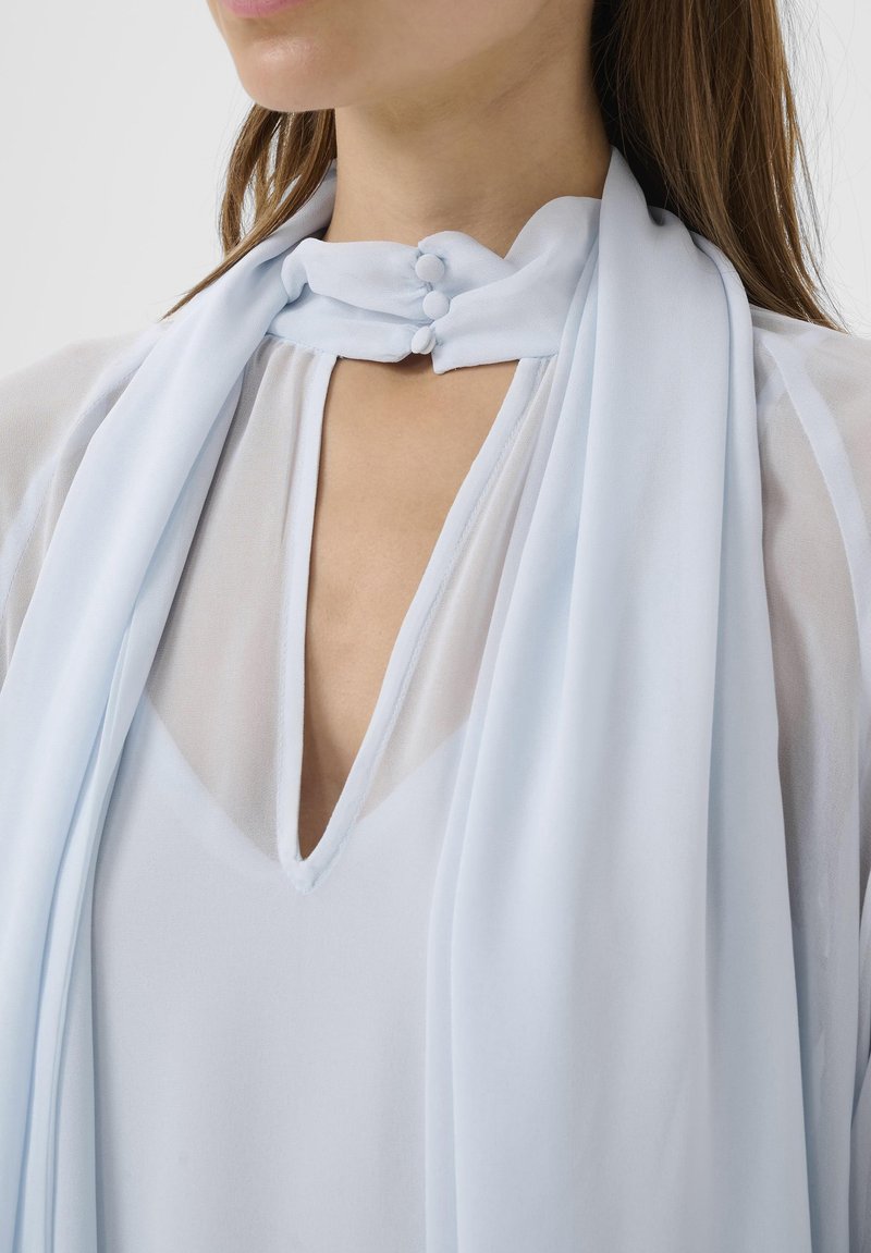 Blouse légère bleu clair en tissu transparent avec un col en V profond et un col montant fermé par trois boutons recouverts de tissu.
