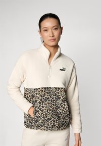 Cremefarbener Fleece-Pullover mit Leopardenmuster im unteren Bereich, halber Reißverschluss vorne und schwarzem Puma-Logo auf der Brust. Weicher Stoff, lässiger Schnitt.