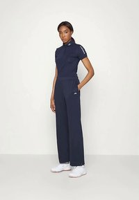 Mulher em pé, vestindo uma camisola de polo azul marinho de mangas curtas e calças de perna larga com sapatos esportivos brancos, contra um fundo claro e simples.