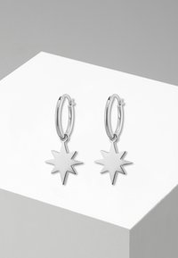 Orecchini a cerchio in argento con pendente a forma di stella a otto punte, superficie liscia, finitura lucida e design minimalista.