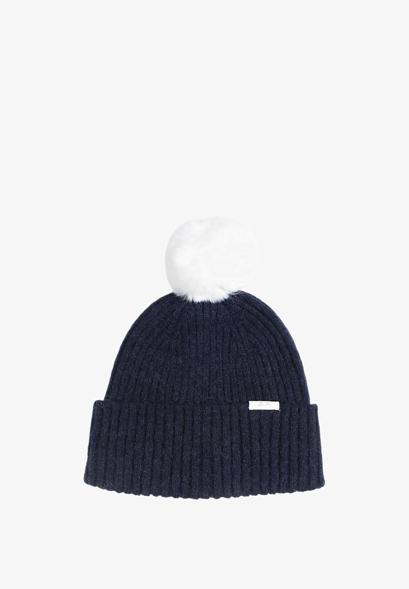 Beanie lavorato a maglia di colore blu navy con texture a coste, abbellito da un pom-pom in pelliccia sintetica bianca. Presenta un piccolo etichetta con logo bianco sul bordo.