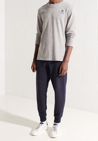 Eine Person steht gegen eine weiße Wand und trägt ein graues Langarmshirt, marineblaue Jogginghosen und weiße Turnschuhe, die Hände entspannt an den Seiten.