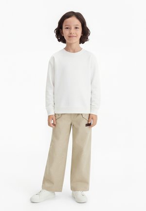 Bambino con capelli scuri indossa una felpa bianca, pantaloni beige a gamba larga con piccole macchie bianche e scarpe da ginnastica bianche, in piedi contro uno sfondo bianco.