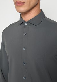 Grijs shirt met lange mouwen en een spitskraag, voorzien van metalen knopen en een textuurrijke stof. Close-up die de stiksel details toont.