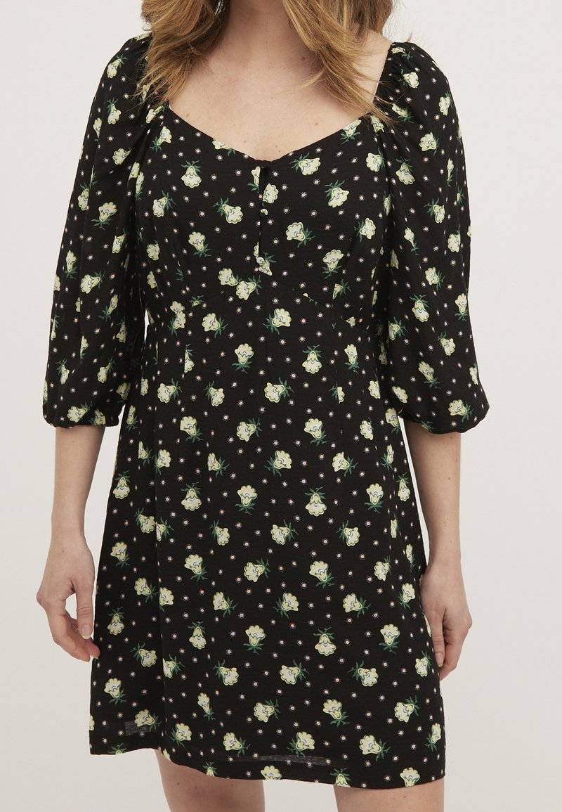 Femme portant une robe noire à imprimé floral blanc avec des manches trois-quarts bouffantes sur un fond uni.