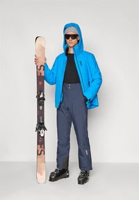 Blå vadderad skidjacka med huva, svart baslager, marinblå skidbyxor och svarta skidpjäxor, håller i skidor med bindningar, mot en neutral bakgrund.