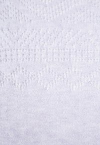 Vitt stickat tyg med ett strukturerat mönster av små upphöjda prickar och zickzackdesign, vilket skapar en mjuk och detaljerad yta.