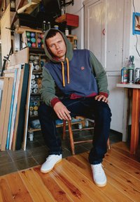 Kapuzensweatshirt mit Farbblöcken in Marineblau, Grün und Burgunderrot, mit orangefarbenen Kordeln. Dunkle Jeans und weiße Sneakers.