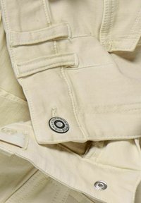 Jasnobeżowa tkanina z dwoma prostokątnymi paskami, srebrnym okuciem i guzikem z wygrawerowanym tekstem. Gładka struktura, starannie dopasowane detale.