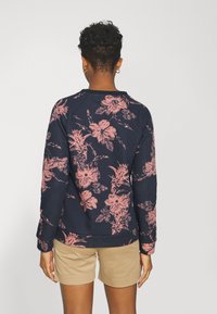 Une personne aux cheveux courts et bouclés porte un sweat-shirt bleu marine avec des motifs floraux roses et un short beige, se tenant de dos contre un fond uni.