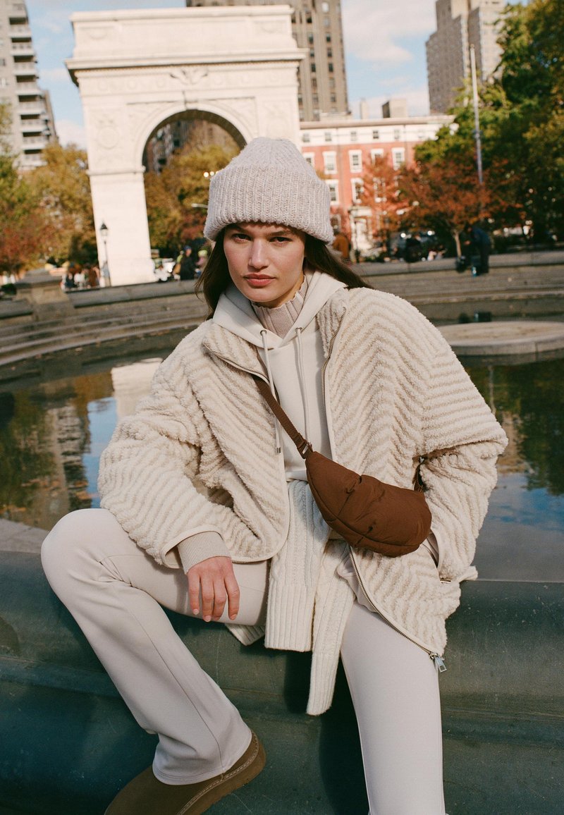 Giacca in pile beige a coste sopra una felpa leggera, abbinata a pantaloni color crema e stivali marroni. Una borsa a tracolla marrone è indossata a tracolla.