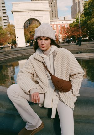 Beige, geribbelde fleecejas over een lichte hoodie, gecombineerd met crèmekleurige broek en bruine laarzen. Een bruine tas wordt schuin over het lichaam gedragen.