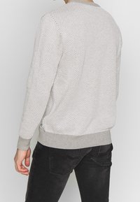 Mężczyzna w jasnoszarym swetrze o fakturze i czarnych dżinsach pokazany od tyłu na tle jednolitego koloru.