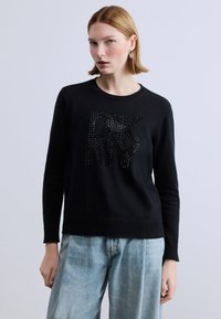 Maglione nero con scollo e orlo a coste, decorato con la scritta "DKNY" con borchie. Abbinato a jeans a gamba larga azzurri.