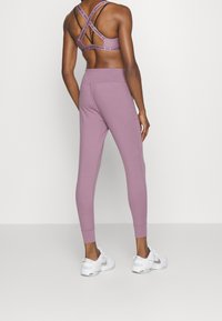 Jasnofioletowe legginsy do treningu i pasujący stanik sportowy mają krzyżujące się ramiączka, gładką teksturę i dopasowany krój. Sneakersy są białe z srebrnymi akcentami.