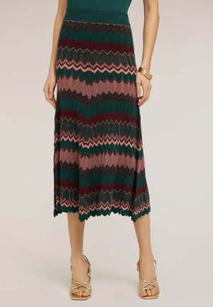 Kobieta nosząca plisowaną spódnicę midi w zielone, bordowe i różowe zygzakowate wzory, do tego złote sandały na paski i bransoletę z koralików.